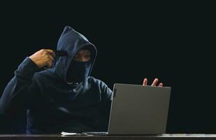 Portrait hacker spy man one person in black hoodie sitting on table looking computer laptop used login password attack security to data digital internet network system night dark background copy space photo