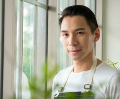 Portrait garden handsome cool young Asian man wearing white blouse with long black hair and smile fresh happy with bright smile look hand holding pot small tree leaf green plant in room shop and relax photo