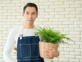 Portrait garden handsome cool young Asian man wearing white blouse with long black hair and smile fresh happy with bright smile look hand holding pot small tree leaf green plant in room shop and relax photo