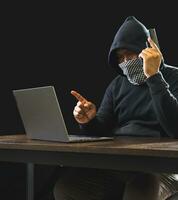 Portrait hacker spy man one person in black hoodie sitting on table looking computer laptop used login password attack security to data digital internet network system night dark background copy space photo