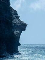 Majestic cliff face overlooking a serene ocean, with waves gently crashing at its base under a soft blue sky. photo