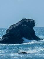 Isolated rock formation in the sea with waves crashing around it under a clear blue sky. photo