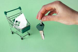 A hand holds the keys and next to it is a mini-trolley with a house made of white paper. photo