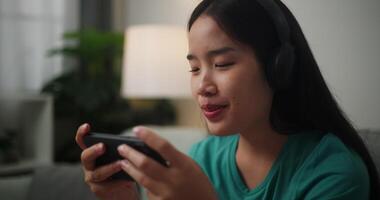 beeldmateriaal van jong Aziatisch vrouw vervelend hoofdtelefoons geniet spelen online esport spellen Aan smartphone zittend Aan sofa in de leven kamer Bij thuis, gamer levensstijl concept. video