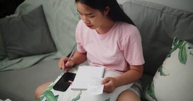 Aufnahmen von jung asiatisch Frau prüft ihr Finanzen mit ein Taschenrechner auf Smartphone und macht Anmerkungen im ein Notizbuch während Sitzung auf das Sofa beim Zu Hause zu Hause Finanzen video