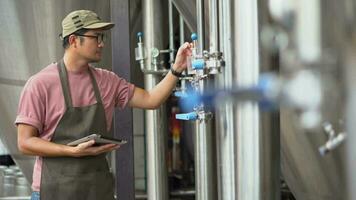 Asia male brewery worker with digital tablet in hands setting parameters on storage tank producing beer at factory. video