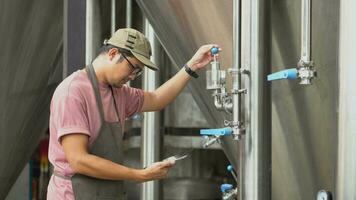 Asia male brewery worker with digital tablet in hands setting parameters on storage tank producing beer at factory. video