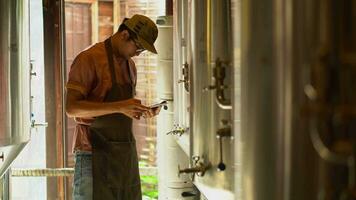 Asia male brewery worker with digital tablet in hands setting parameters on storage tank producing beer at factory. video