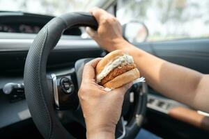 Asian lady holding hamburger to eat in car, dangerous and risk an accident. photo