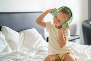 Little girl emotional enjoying music using green kids headphones in home bed. High quality photo