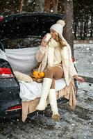 joven mujer en de lana sombrero se sienta en maletero de coche. niña tener un divertido en el coche. invierno viaje en Nevado clima. alto calidad foto