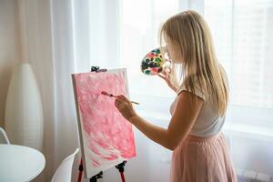 Young blonde woman artist with palette and brush painting abstract pink picture on canvas near window. Art and creativity concept. High quality photo