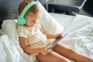 Little girl in headphones using digital tablet while listening to music or playing game in home bed. High quality photo