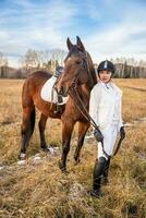 Beautiful blond professional female jockey standing near brown horse in field in winter. Friendship with horse. High quality photo