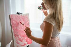 Young blonde woman artist with palette and brush painting abstract pink picture on canvas at home. Art and creativity concept. High quality photo