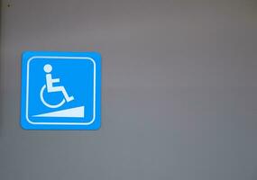 White sign of handicapped ramp for wheelchair on bright blue square shape on gray wall of building. photo