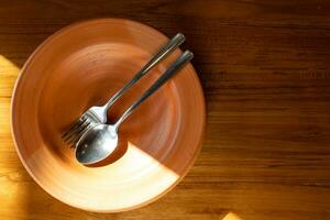 Close up stainless steel cutlery on clay plate. Clay Plate with spoon and fork on wooden table with sunlight. photo