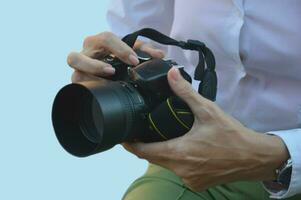 A professional photo camera in the hands of a woman in a white shirt on a blue background. A girl photographer holds a camera in her hand.