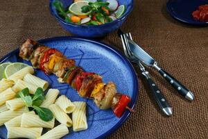 Grilled meat skewers with pasta on blue plate with vegetable salad photo