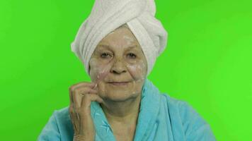 mayor abuela en bata de baño. antiguo mujer aplicando hidratante crema en cara video