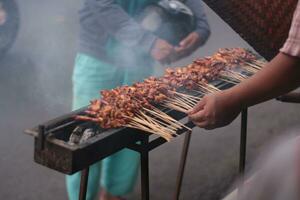 Chicken satay over traditional charcoal fire. The satay burns with smoke and an appetizing appearance. hands cooking satay on the grill. people are already queuing. photo