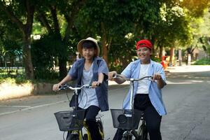 dos asiático Niños quien son amigos paseo bicicletas en el local parque en día festivo. ellos son ambos contento y teniendo divertida. fiesta actividad concepto. suave y selectivo enfocar. foto