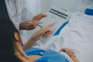 The senior woman specialist doctor with stethoscope checking up happy young Asian female patient lying in bed with receiving an intravenous saline drip in recovery room. Healthcare, medical insurance. photo