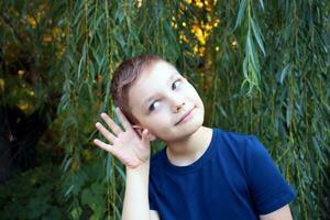 Portrait of a 9 year old boy on a background of leaves. A handsome boy in a blue t-shirt holds his hand near his ear. photo