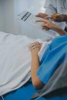 The senior woman specialist doctor with stethoscope checking up happy young Asian female patient lying in bed with receiving an intravenous saline drip in recovery room. Healthcare, medical insurance. photo