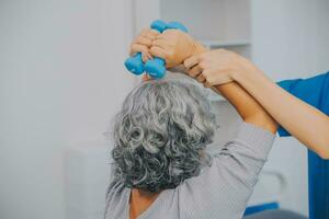 Old woman training with physiotherapist using dumbbells at home. Therapist assisting senior woman with exercises in nursing home. Elderly patient using dumbbells with outstretched arms. photo