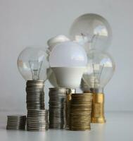 LED lamp and filament bulbs behind the piles of coins isolated on white. Concept photo for metaphoric increasing of energy saving