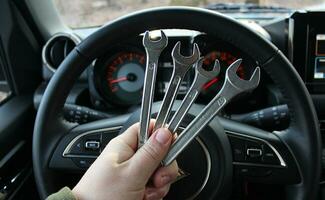 A mechanic holds a bunch of variety diameter wrenches in front of the steering wheel and blurred dashboard in car with the ignition on photo