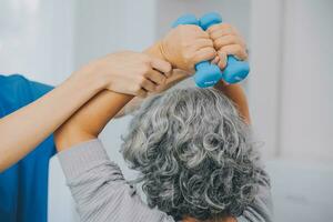 Old woman training with physiotherapist using dumbbells at home. Therapist assisting senior woman with exercises in nursing home. Elderly patient using dumbbells with outstretched arms. photo