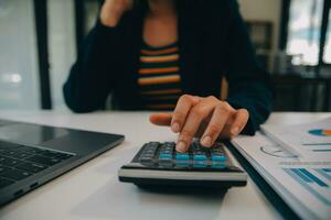 encantadora joven mujer de negocios asiática sentada en una computadora portátil en la oficina, haciendo un informe que calcula el documento de verificación del servicio de ingresos internos del saldo. foto