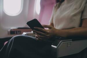 Asian woman passenger sitting in airplane near window and reading news from social networks or using travel applications in smartphone photo