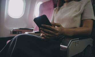 Asian woman passenger sitting in airplane near window and reading news from social networks or using travel applications in smartphone photo