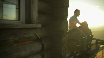 Young man sit near old wooden house. Sun beams. Slow motion video