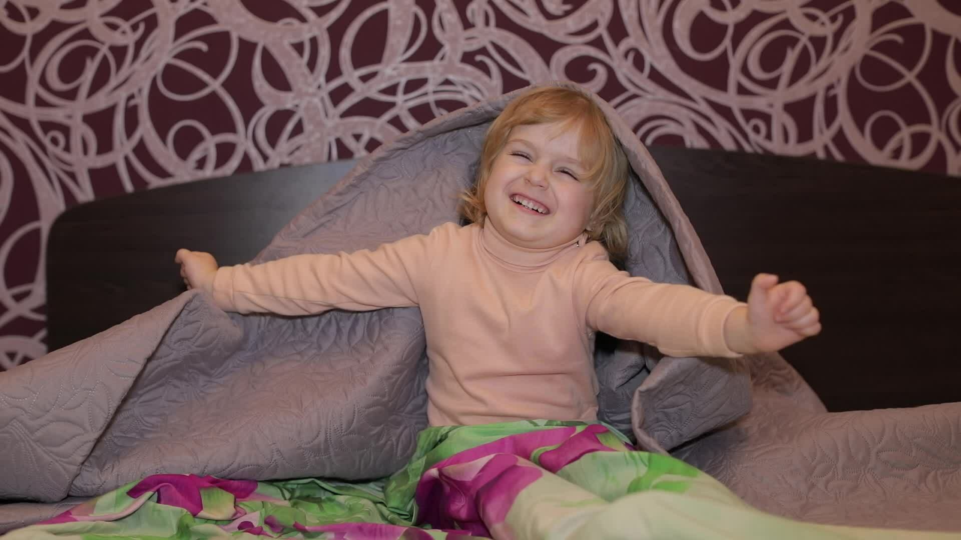 Little cheerful girl hides under blanket and watching TV. Concept of