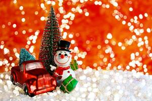 On a red background with bokeh, a cheerful snowman near a pickup truck with a Christmas tree. Christmas banner, card with place for text photo