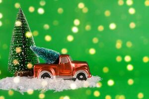 A red pickup truck is carrying a Christmas tree with artificial snow on a green background with bokeh. Banner, New Year card with copy space photo