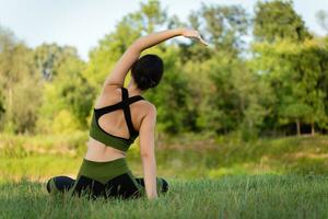 View from the back. Sitting on the grass, a teenager girl performs tilts to the sides with a beautiful view of the summer nature. Physical form, harmony photo