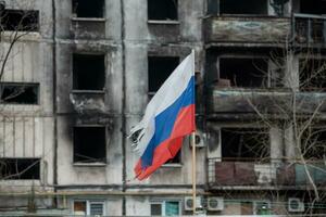 tricolor flag of Russia against the background of destroyed and burnt houses in Ukraine photo