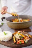 Dish named Lomo saltado served on a wooden board and in the background some hands serving from a pot. photo