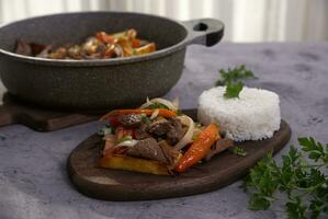 Dish named Lomo saltado served on a wooden board and in the background some hands serving from a pot. photo
