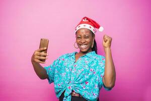 overexcited black african female millenial celebrating with her smart phone after receiving good news. wearing xmas hat standing behind a pink studio wall. christmas concept photo