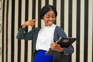 Single happy young executive with long brown hair and black jacket standing in small office holding black binder on her other hand photo