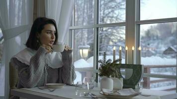 en un invierno mañana, un niña siguiente a ventana bebidas café. nieve fuera de video