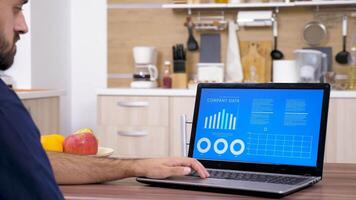 Man looking at animated financial charts and data at the table on his laptop. There is a cozy and modern kitchen in background. Dolly slider 4K footage video