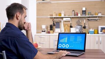 Young businessman at home in the kitchen looking at computer with animated financial charts and data on it video