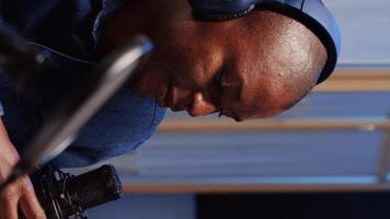 Vertical video African american man doing podcast in studio, wearing headphones and talking on professional microphone with guest. Close up shot of BIPOC content creator interviewing another person